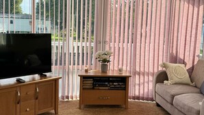 A living room with a large flat-screen TV on a wooden stand, a bookshelf with books, a beige sofa, and a window with vertical blinds. - VALENCIA 2 BEDROOMS LAKESIDE LODGE (Weeley)