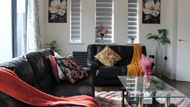 En stue med en sort lædersofa, et glasbord og et rødt pled. Tre vinduer med hvide persienner, en potteplante og en vase med blomster på vindueskarmen.