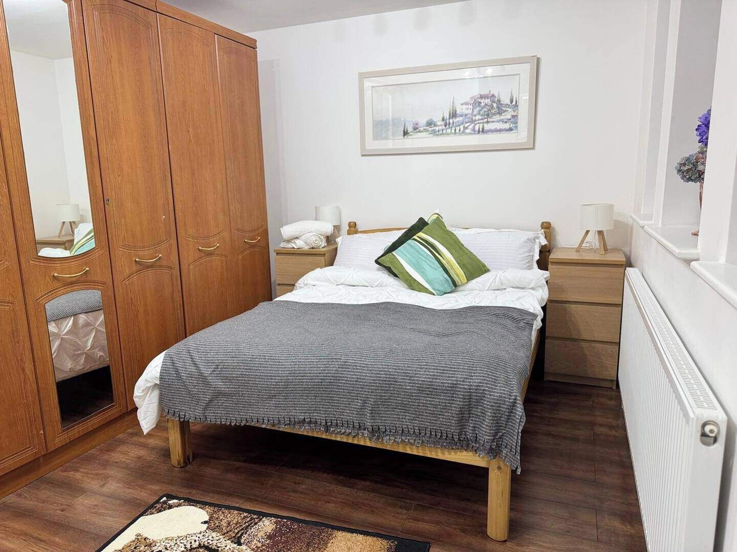 A bedroom with a wooden wardrobe, a bed with white bedding and a grey blanket, a nightstand with a lamp, and a framed picture on the wall.