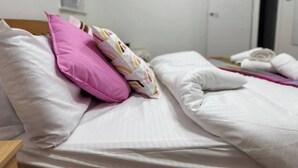 En pænt redt seng med hvide og lyserøde puder, en stribet pude og et hvidt tæppe.