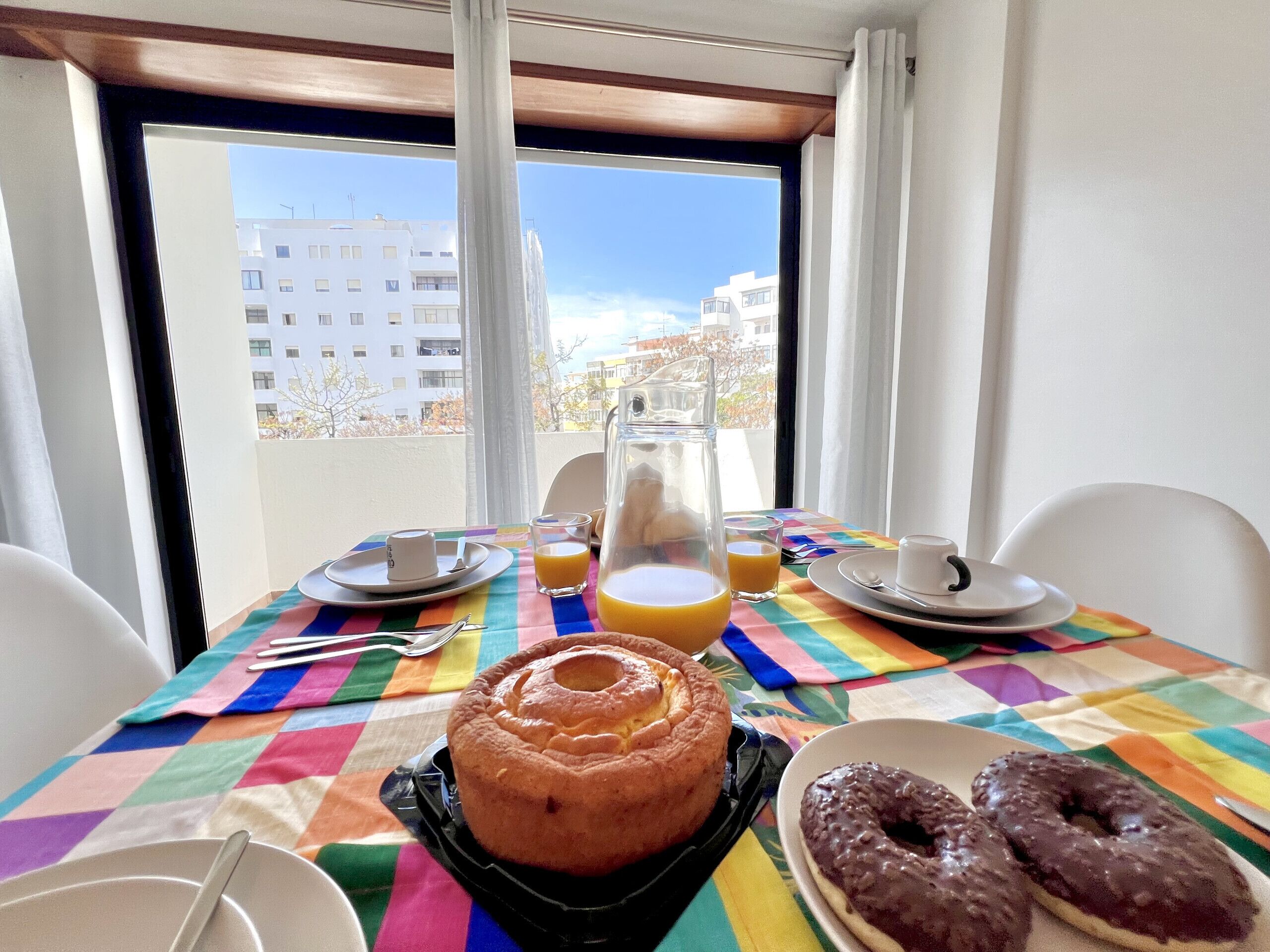 A breakfast table with a colorful tablecloth, a glass pitcher, orange juice, a plate with a pastry, and two chocolate donuts.