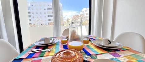 A breakfast table with a colorful tablecloth, a glass pitcher, orange juice, a plate with a pastry, and two chocolate donuts.