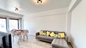 Una sala de estar moderna con un sofá gris, cojines amarillos, una mesa de comedor con sillas y un televisor de pantalla plana.