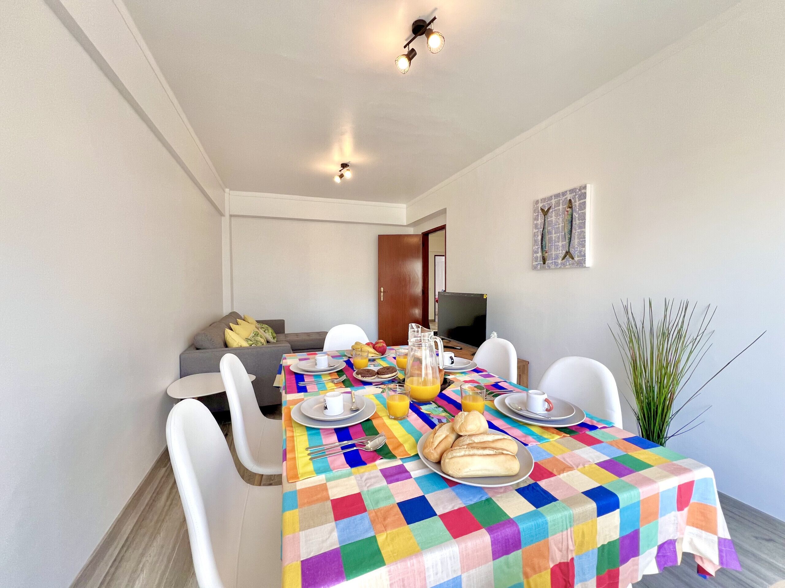 A dining area with a colorful tablecloth, white chairs, and a television in the background.