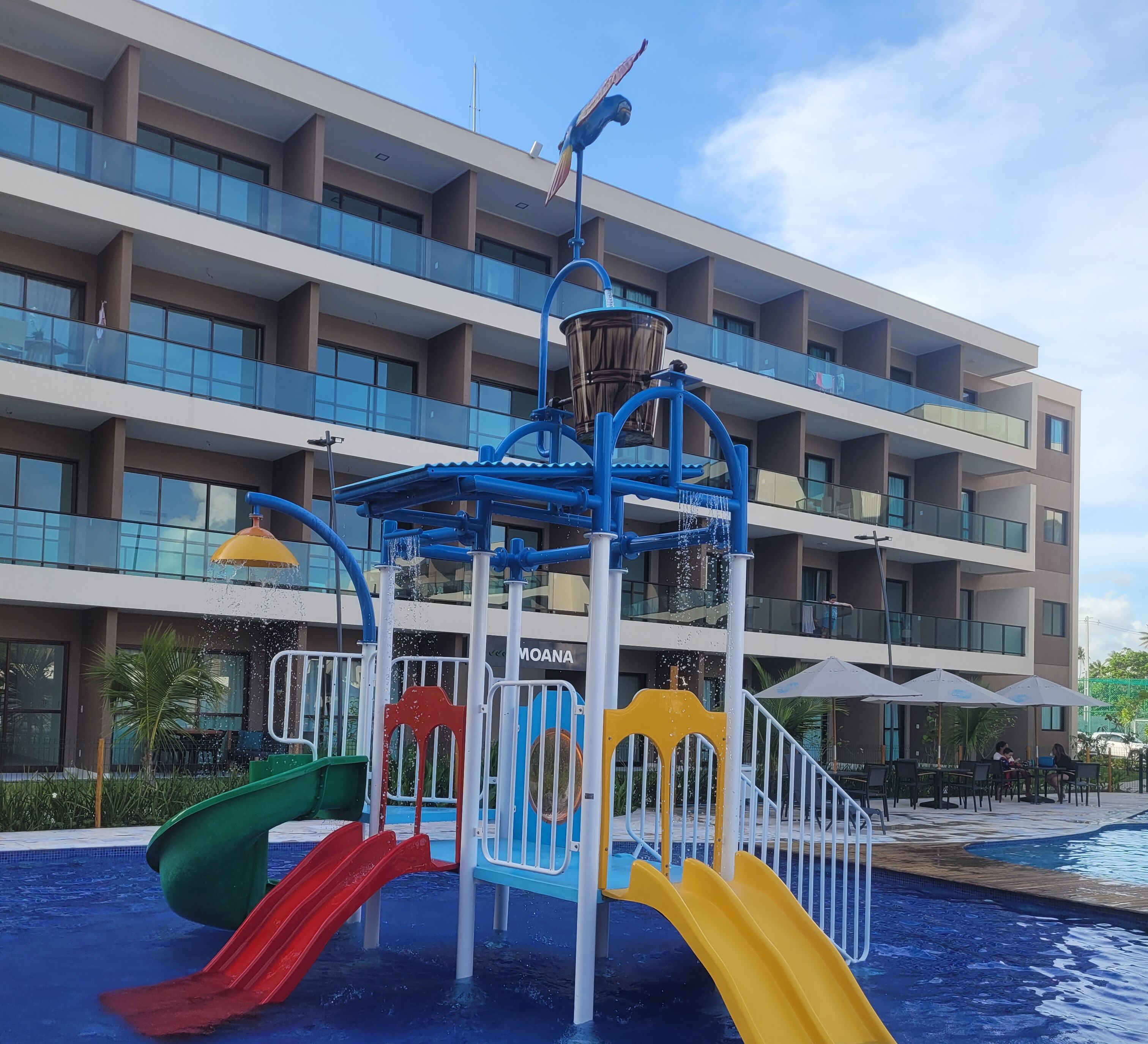 Uma área de recreação aquática com escorregadores e uma fonte, em frente a um hotel com várias sacadas.