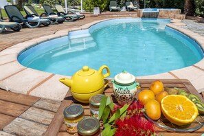 Un espace aménagé au bord de la piscine, avec des chaises longues, une théière jaune, un assortiment de fruits et des pots de confiture.