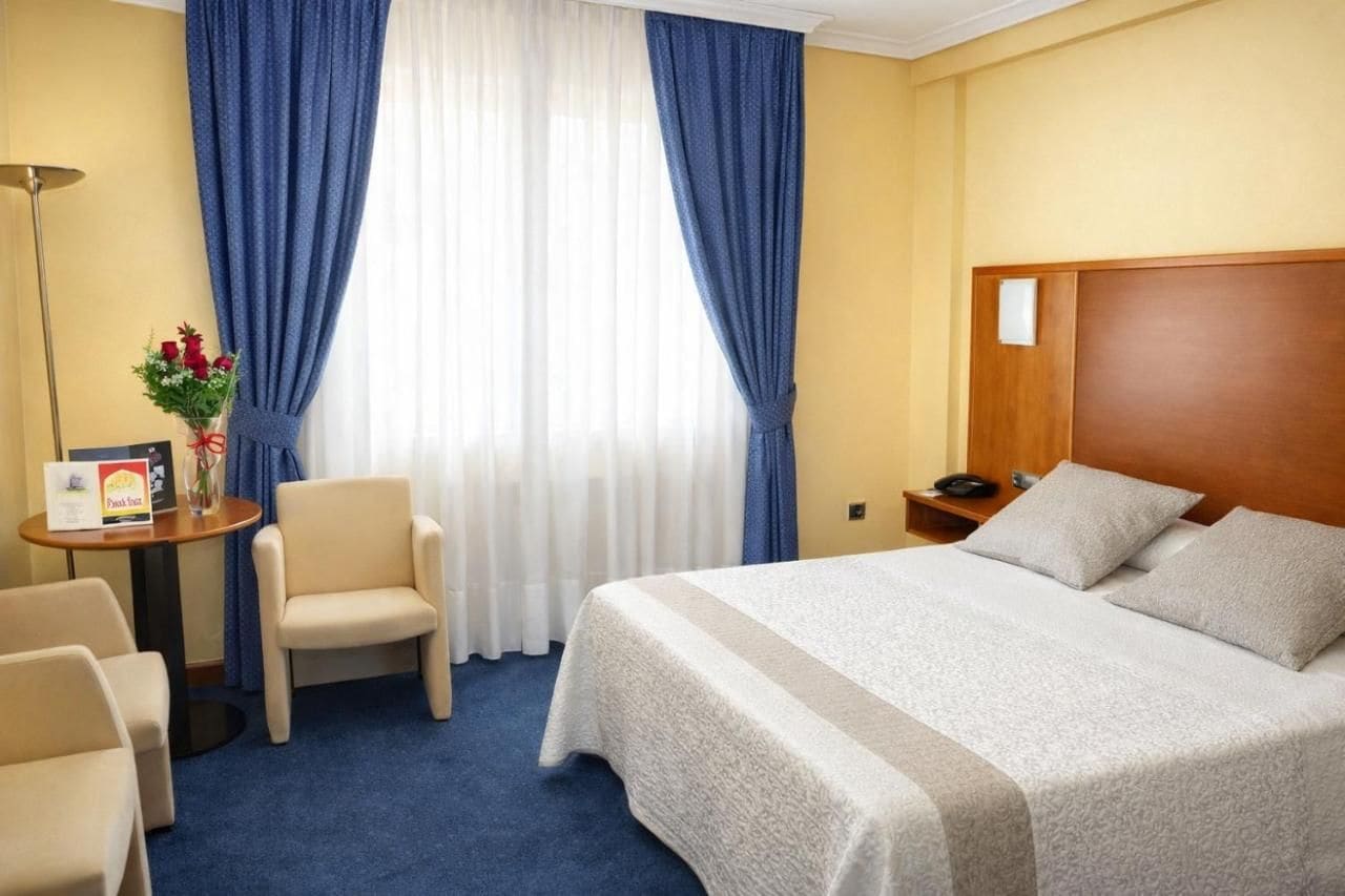Habitación de hotel con una cama grande, una silla, una mesita con un jarrón de flores y una mesita de noche con un teléfono.