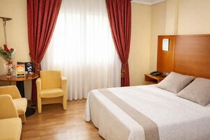 Habitación de hotel con una cama grande, un escritorio con teléfono, una silla y un ventanal con cortinas.
