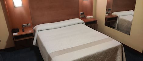Habitación de hotel con cama, mesita de noche, espejo y luz empotrada en la pared.