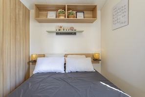 A bedroom with a bed, two bedside tables with lamps, a shelf above the bed with decorative items, and a wall with a framed message. - Mobile Home 'Family's Dream P16 La Marina' with Shared Pool, Private Terrace and Air Conditioning (Canet-en-Roussillon)