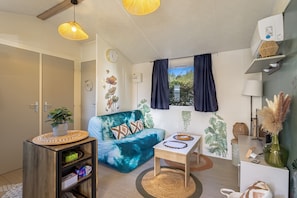 A living room with a sofa, a small table, a plant, and a window with curtains. - Mobile Home 'Family's Dream P16 La Marina' with Shared Pool, Private Terrace and Air Conditioning (Canet-en-Roussillon)