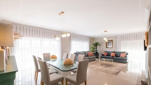 Uma sala de estar moderna com uma mesa de jantar de vidro, sofás cinzas e uma janela grande com cortinas leves.