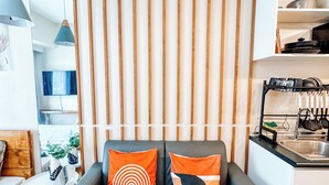 A modern living room with a grey sofa, two orange and black patterned cushions, a wooden slat wall, a small white table with a vase, and a kitchen area with a sink and utensils. - A Modern Zen Hideaway (Iloilo)