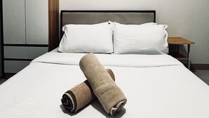 Un lit bien fait, avec du linge de lit blanc et une serviette enroulée, une table de chevet en bois et une armoire.