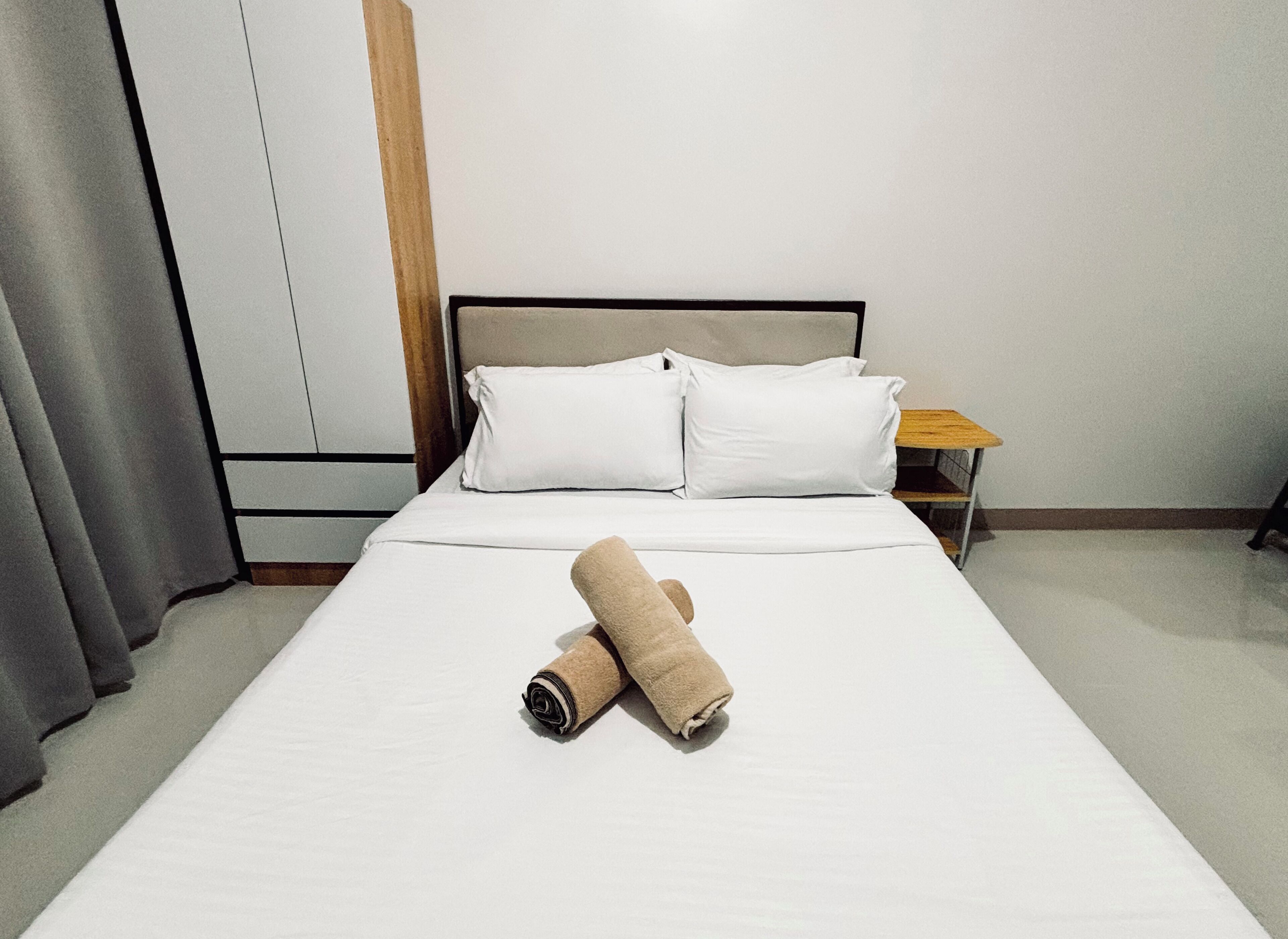 A neatly made bed with white linens and two rolled towels, a wooden nightstand with a lamp, and a grey curtain.