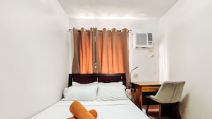 A hotel room with a bed, a desk, a chair, a window with curtains, and an air conditioning unit. - Cozy Family Suite Retreat (Iloilo)