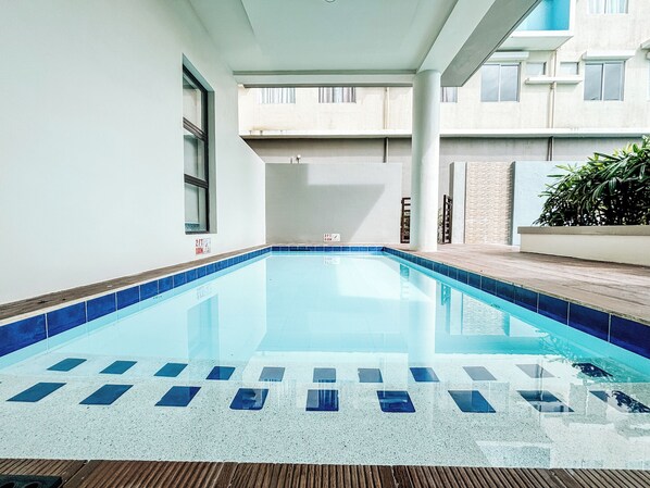 A swimming pool with a tiled edge, a wooden deck, and a building in the background. - Cozy Family Suite Retreat (Iloilo)