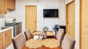 A dining area with a wooden table, four chairs, a TV mounted on the wall, and a refrigerator in the kitchen area. - Cozy Family Suite Retreat (Iloilo)