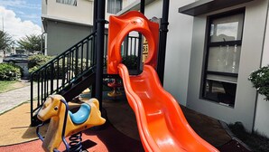 Ein Spielplatz mit Rutsche und einer Pferdebahn, neben einem Gebäude mit Balkonen und Fenstern.