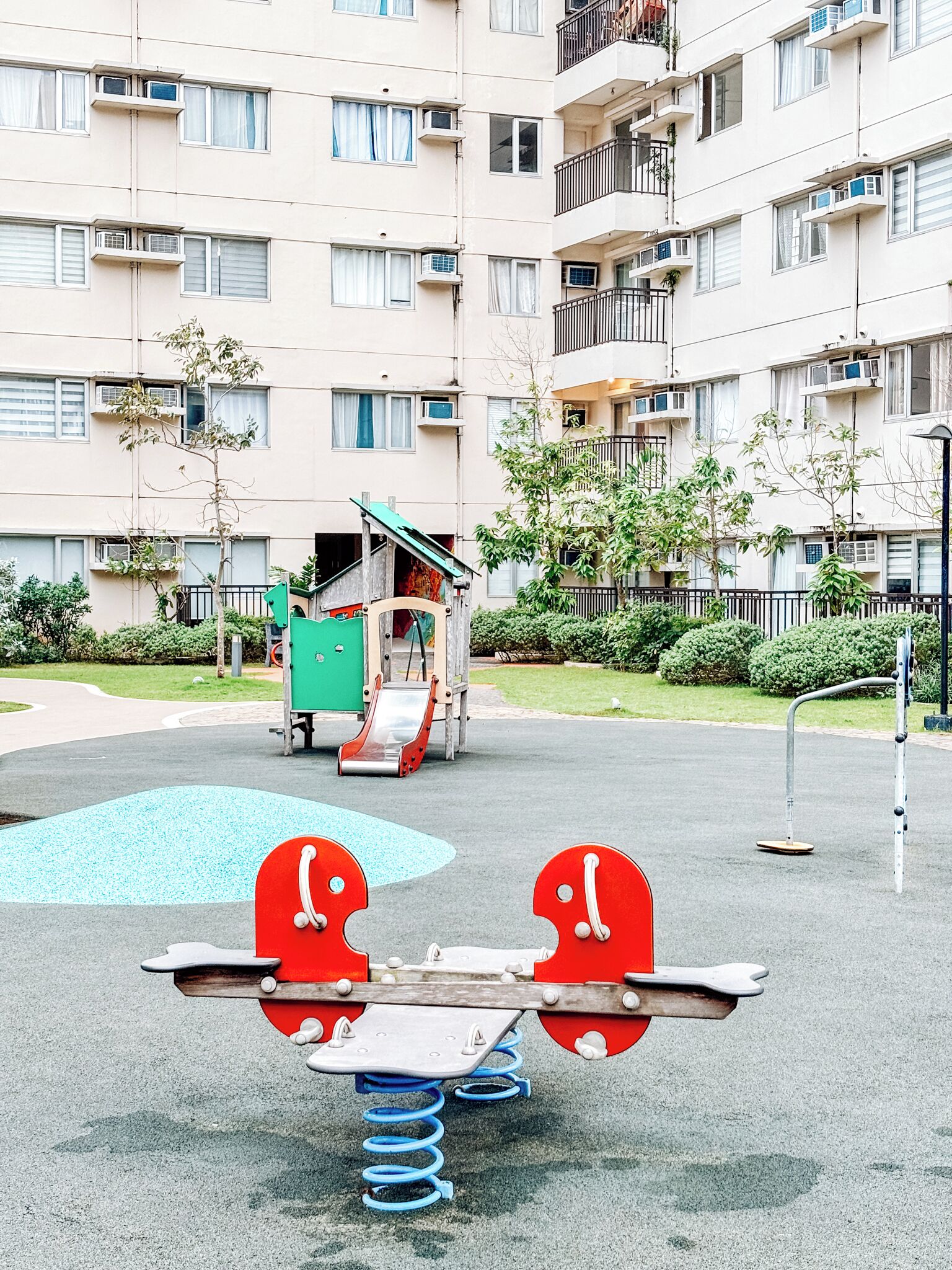 A playground with a seesaw and a slide, surrounded by a residential building with balconies and air conditioning units.