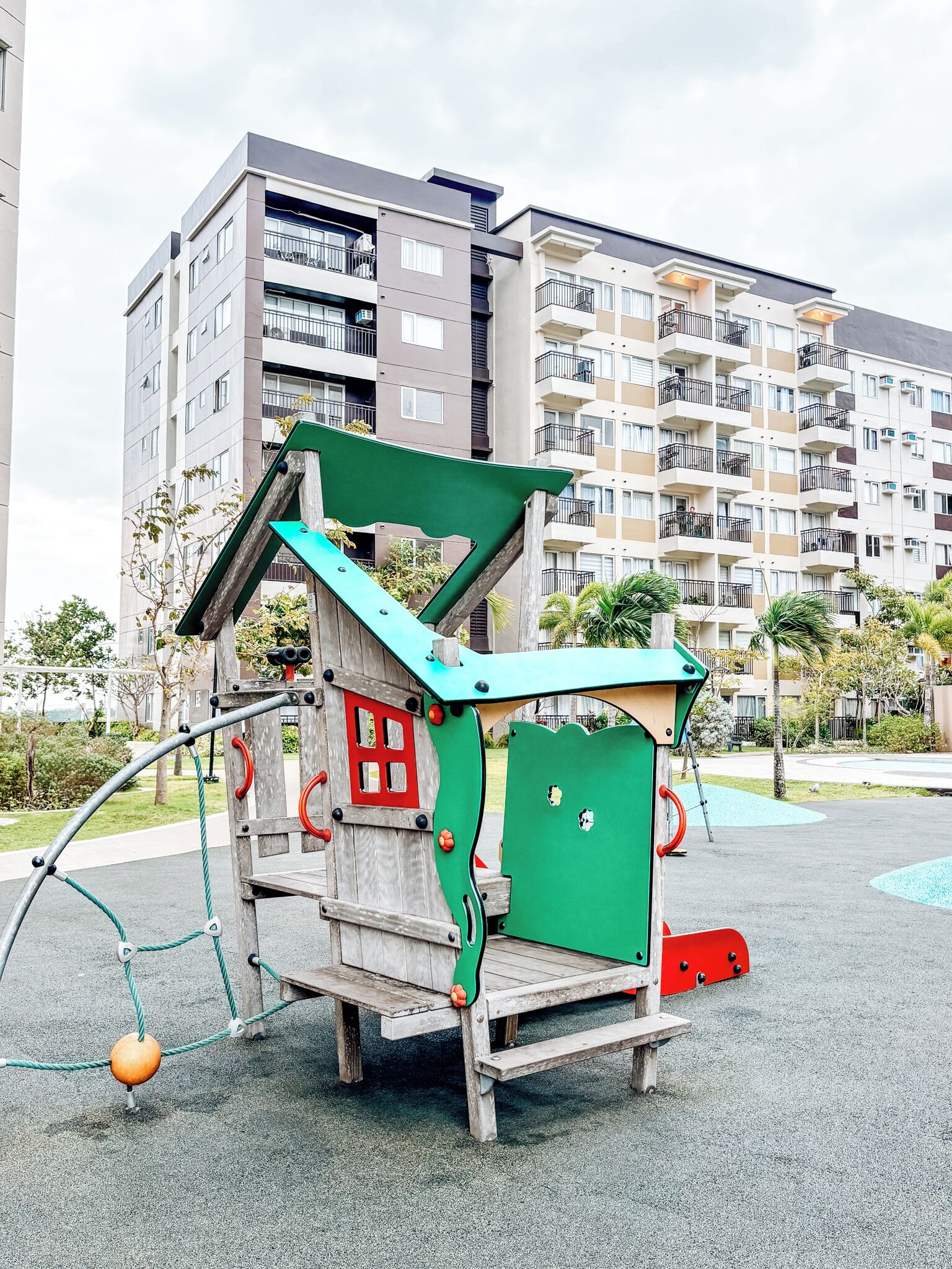 Una estructura de juegos con diseño de casa, rodeada de una zona de estacionamiento y edificios residenciales al fondo.