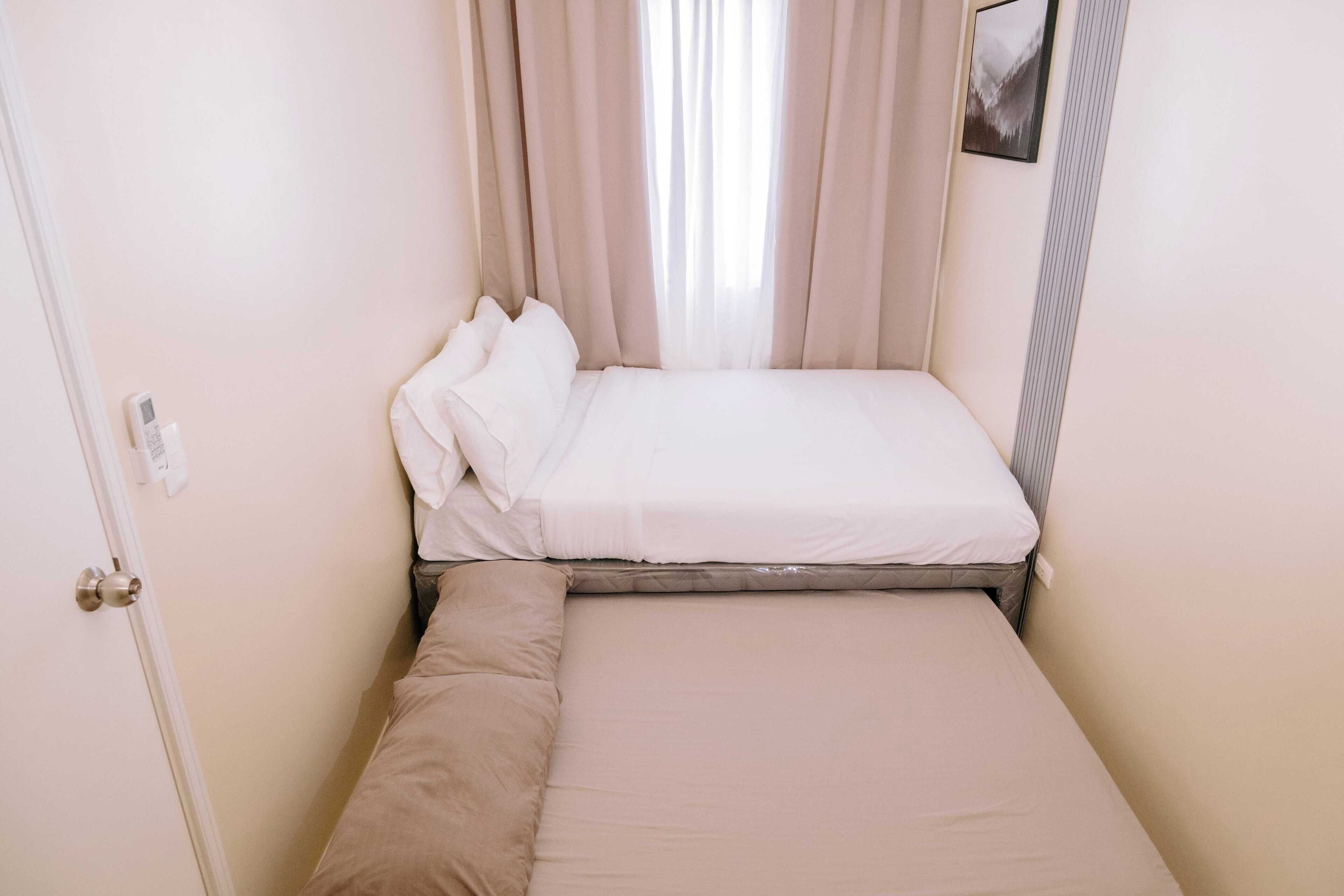A small, single-bed room with a wooden frame, beige bedding, and a window with sheer curtains.