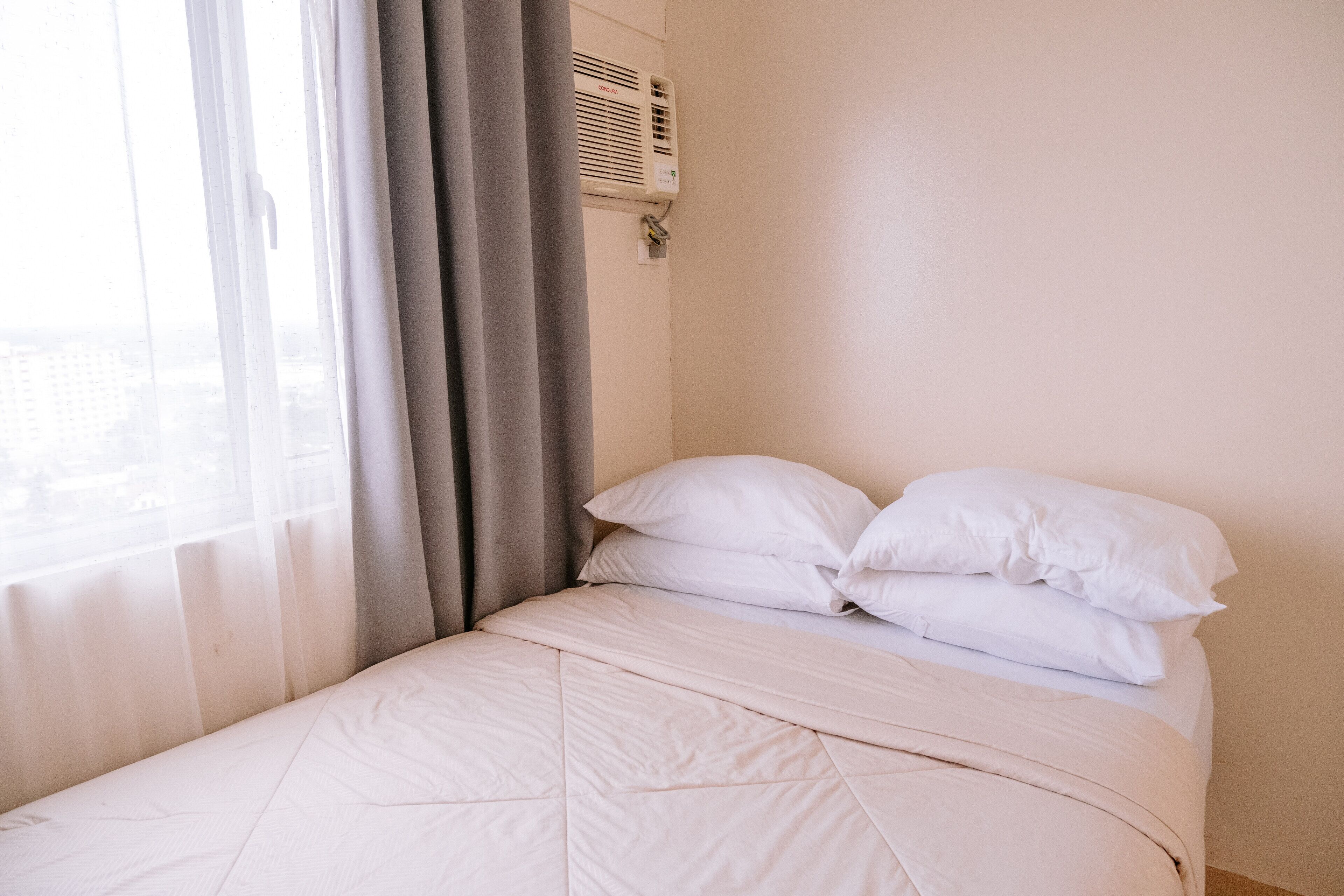 A neatly made bed with white bedding and pillows, a window with sheer curtains, and a wall-mounted air conditioning unit.