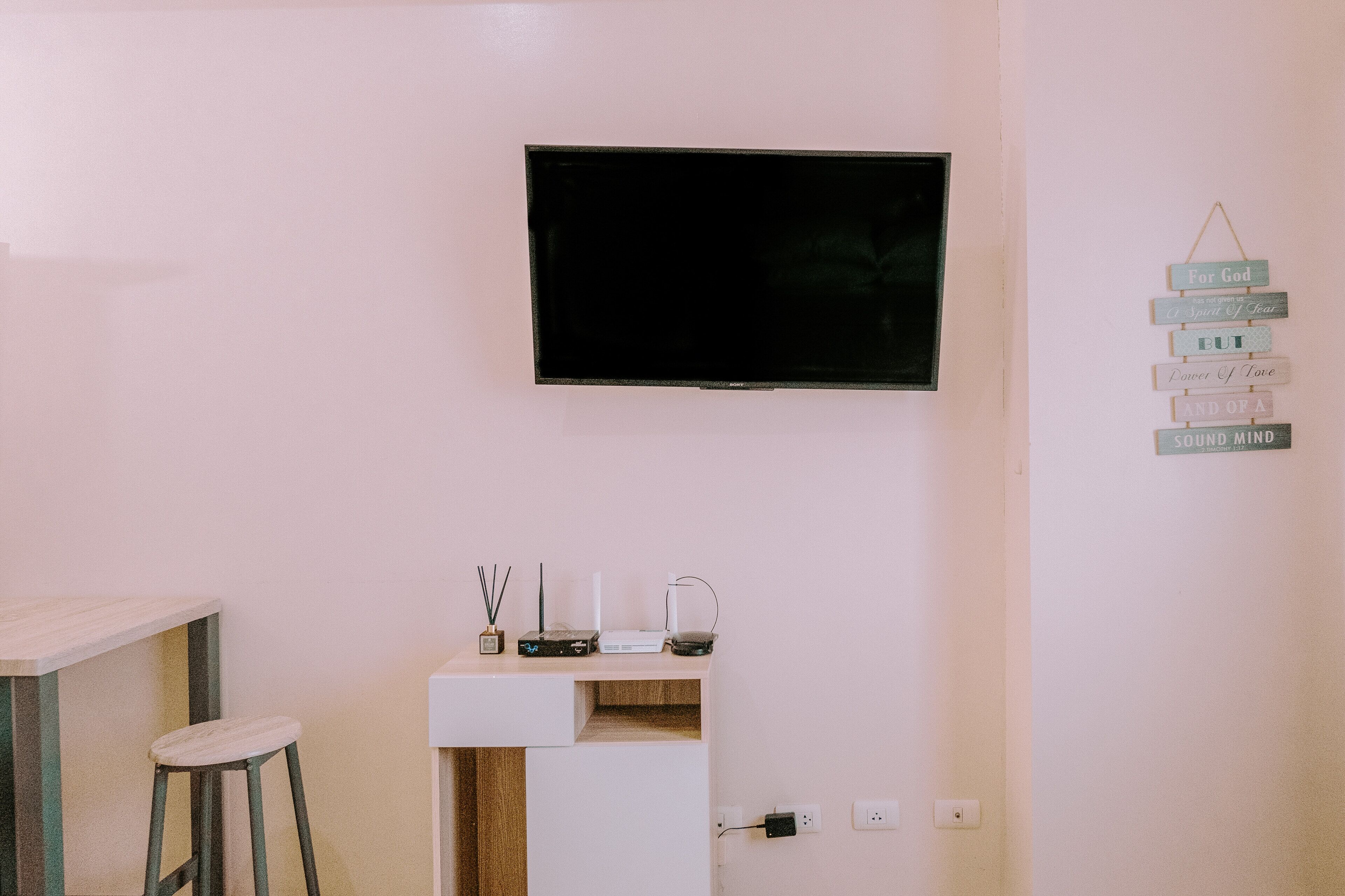 A minimalist room with a flat-screen TV mounted on the wall, a small white desk with a stool, and a wall-mounted sign with inspirational phrases.