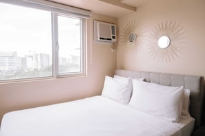 Une chambre avec une grande fenêtre, un lit avec une literie blanche, un climatiseur fixé au mur et deux décorations murales en forme de soleil rayonnant.