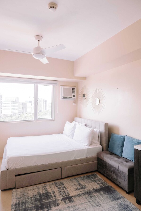 Een slaapkamer met een bed, een plafondventilator, een raam en een aan de muur bevestigde airconditioner.