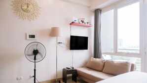 Une chambre d’hôtel équipée d’un lit, d’un ventilateur sur pied, d’un téléviseur fixé au mur, d’un thermostat et d’une fenêtre donnant sur des immeubles.