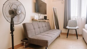 Una sala de estar con un sofá gris, un ventilador de pie, un televisor montado en la pared y una silla.