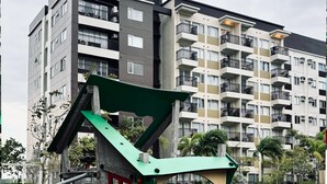 A playground with a green and red play structure, surrounded by a playground area with a pool and a multi-story residential building in the background.