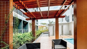 Un patio exterior con mobiliario de mimbre y una mesa de vidrio, que da a una zona de piscina.