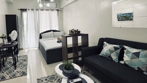 Una habitación de hotel moderna con cama, sofá, mesa de comedor y una mesita de centro. Cuenta con un ventilador de techo, aire acondicionado instalado en la pared y una ventana con cortinas.