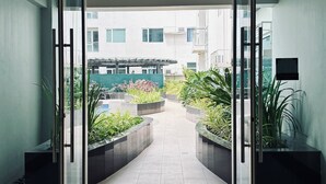 Una entrada de edificio moderna con puertas de vidrio, piso de baldosas y un camino que conduce a un área ajardinada con plantas y una piscina.