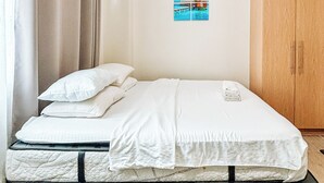 Una cama bien hecha con sábanas blancas, un armario de madera y un cuadro colgado en la pared que representa una escena tropical.