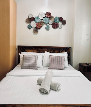 A neatly made bed with white linens and rolled towels, a wooden headboard, and a wall-mounted decorative piece. - Your Peaceful Urban Escape (Iloilo)