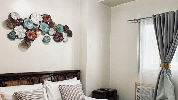 A neatly made bed with white linens and a wooden headboard, a small bedside table, and a wall decoration of variously colored leaves. - Your Peaceful Urban Escape (Iloilo)