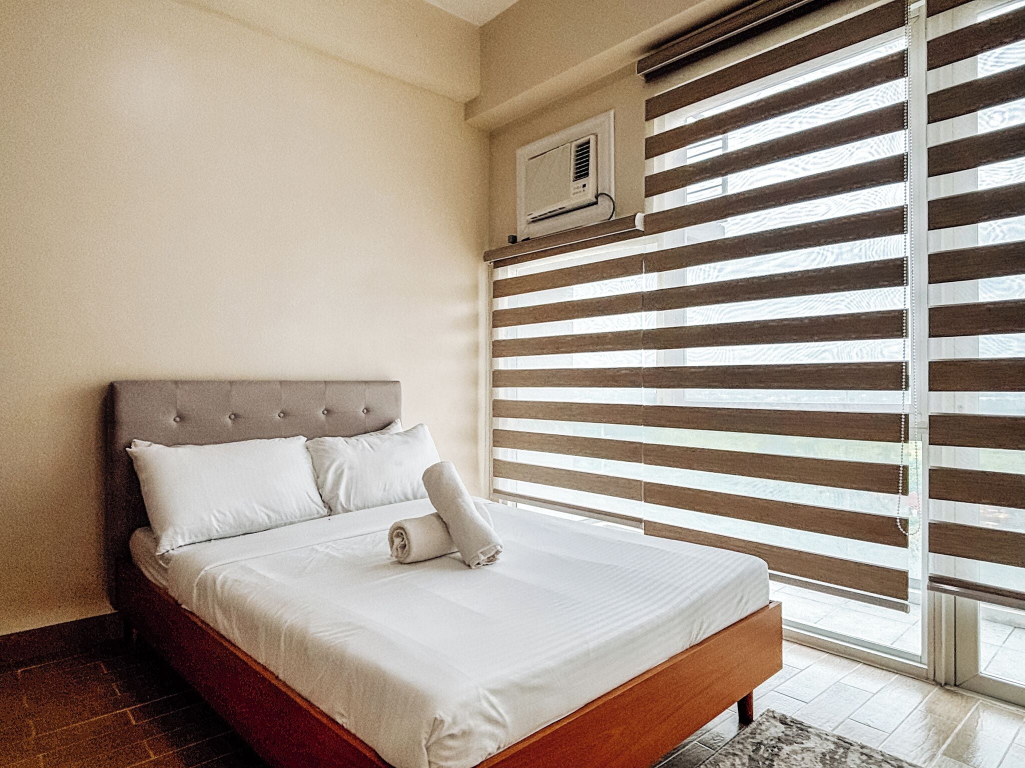 A neatly made bed with white linens and a rolled towel, a wooden headboard, and a window with horizontal blinds.