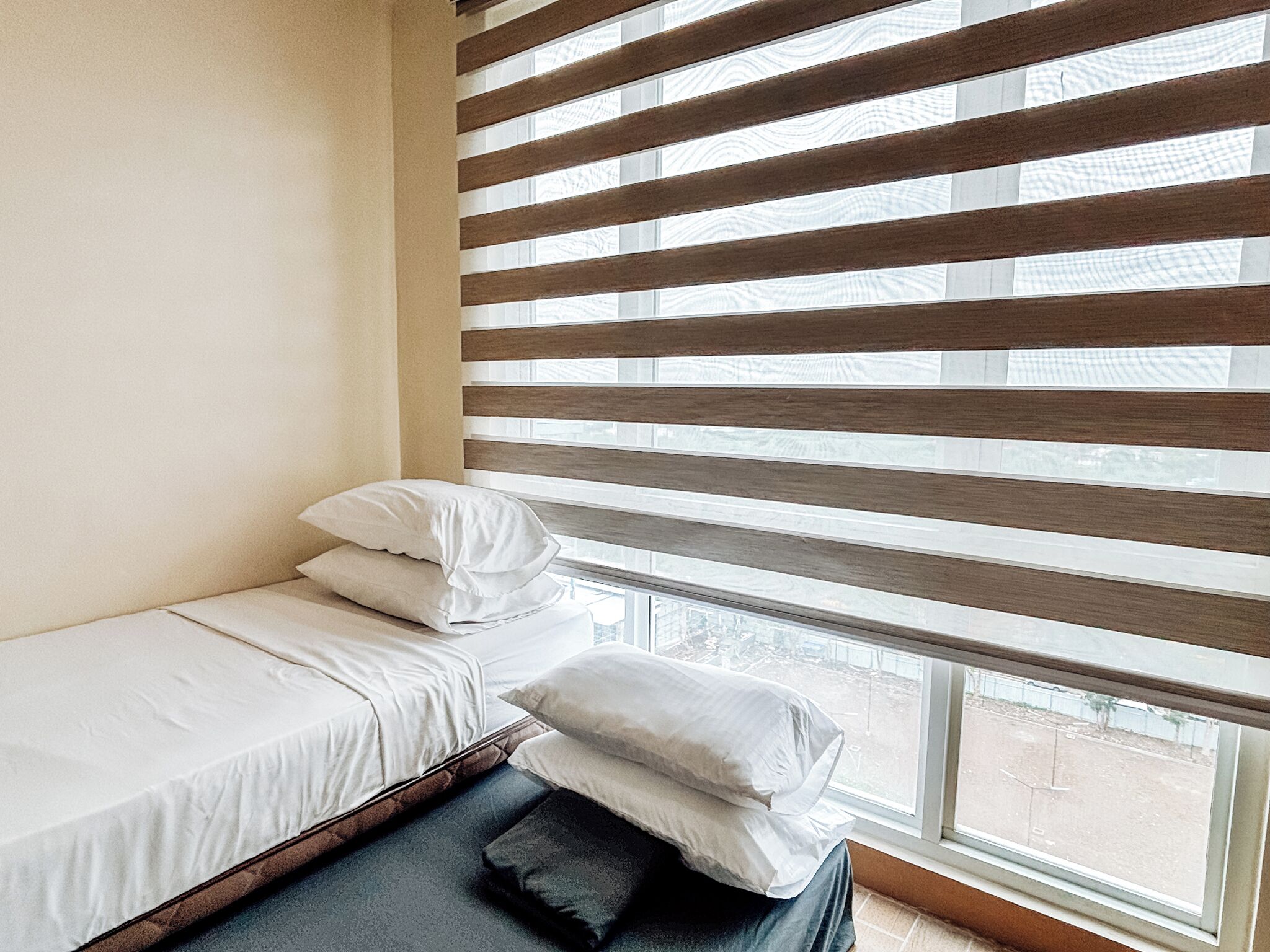 A single bed with white bedding and two pillows, a window with horizontal blinds, and a view of the outside.