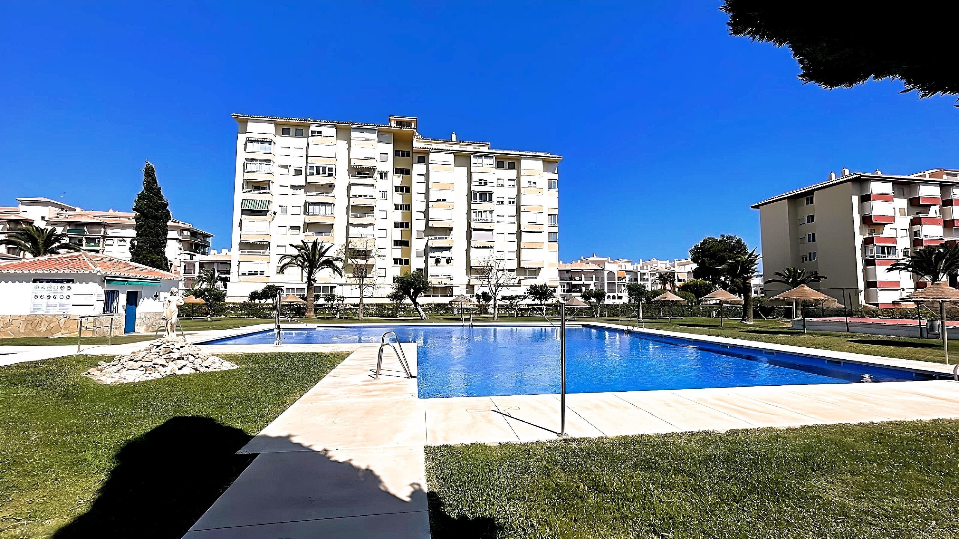 Una piscina con un cielo azul despejado, rodeada de césped y algunas árboles.
