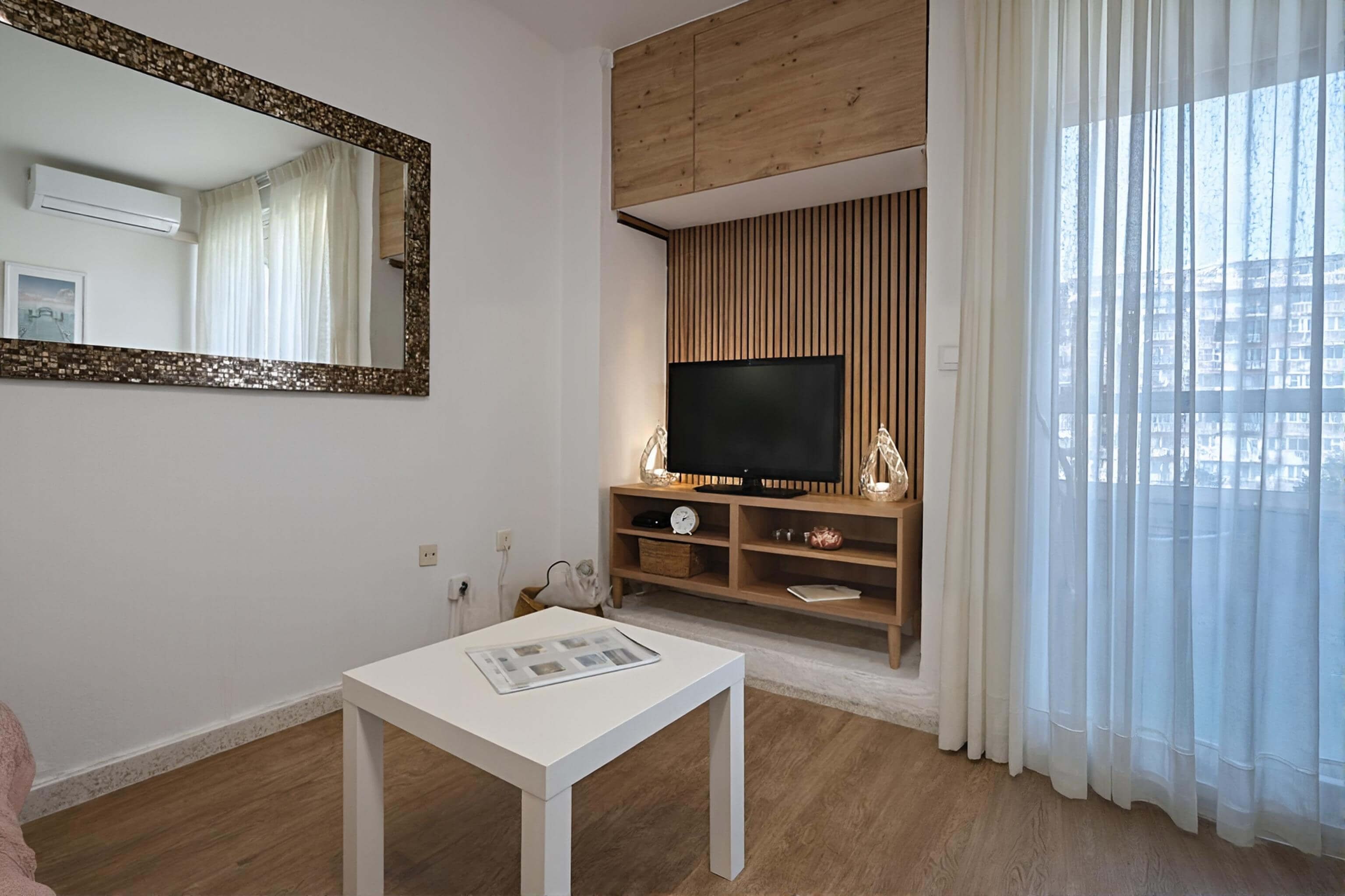 A modern living room with a flat-screen TV, wooden panel wall, white curtains, and a white table with a magazine.