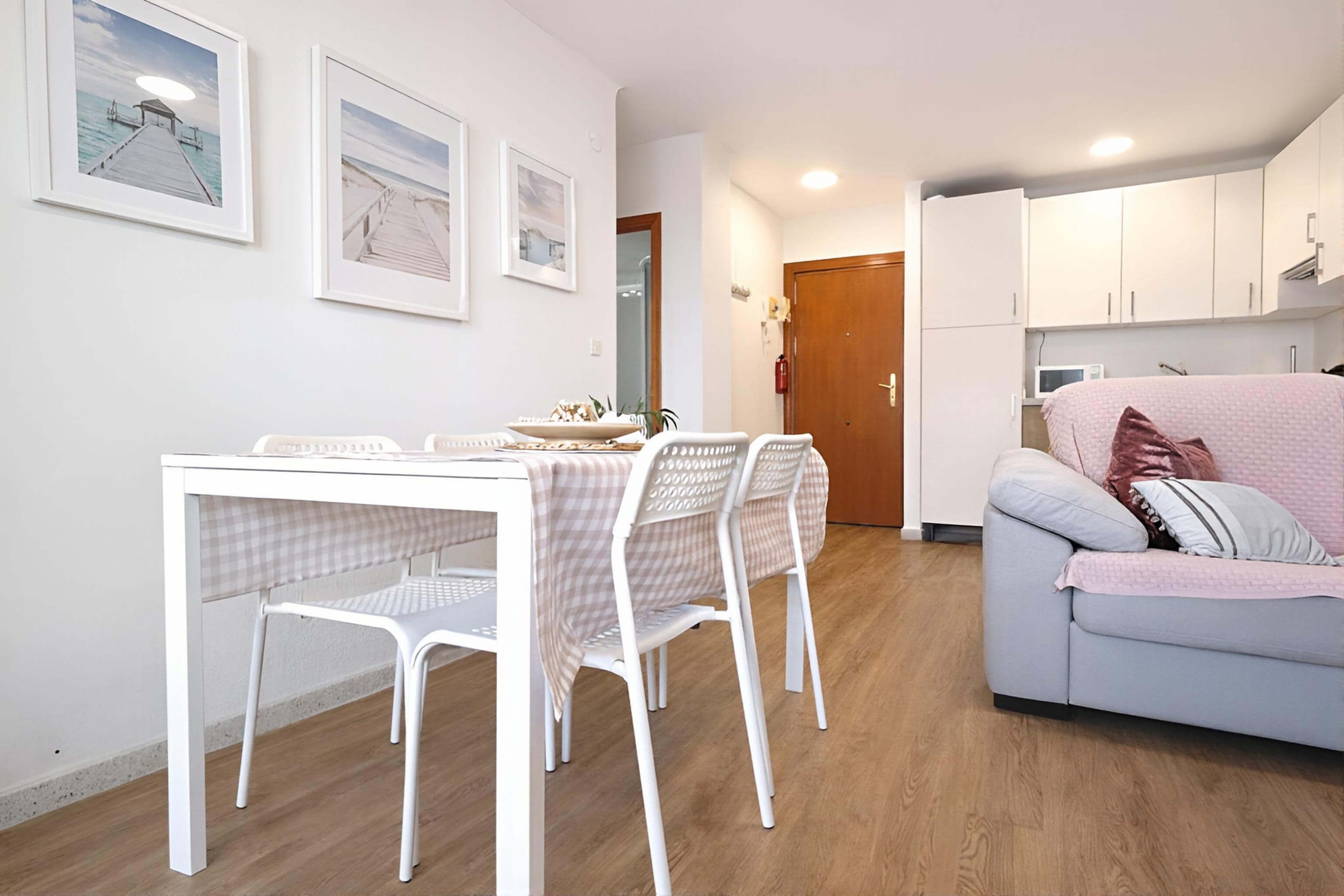 A modern living room with a white sofa, a dining table with checkered tablecloth, and two chairs. There are framed pictures on the wall, a wooden door, and a white cabinet in the background.