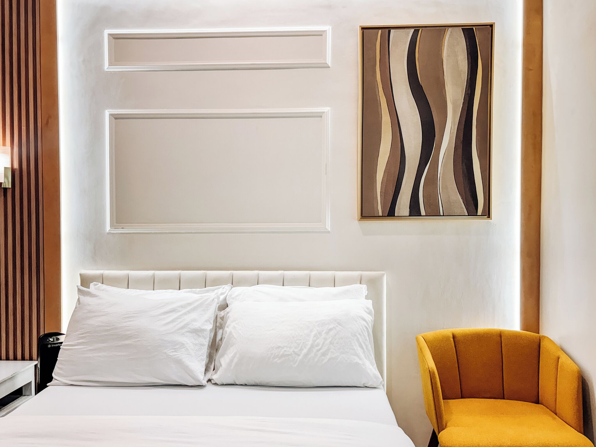 Habitación de hotel con una cama, dos almohadas, cabecero, una obra de arte abstracta colgada en la pared y un sillón amarillo.