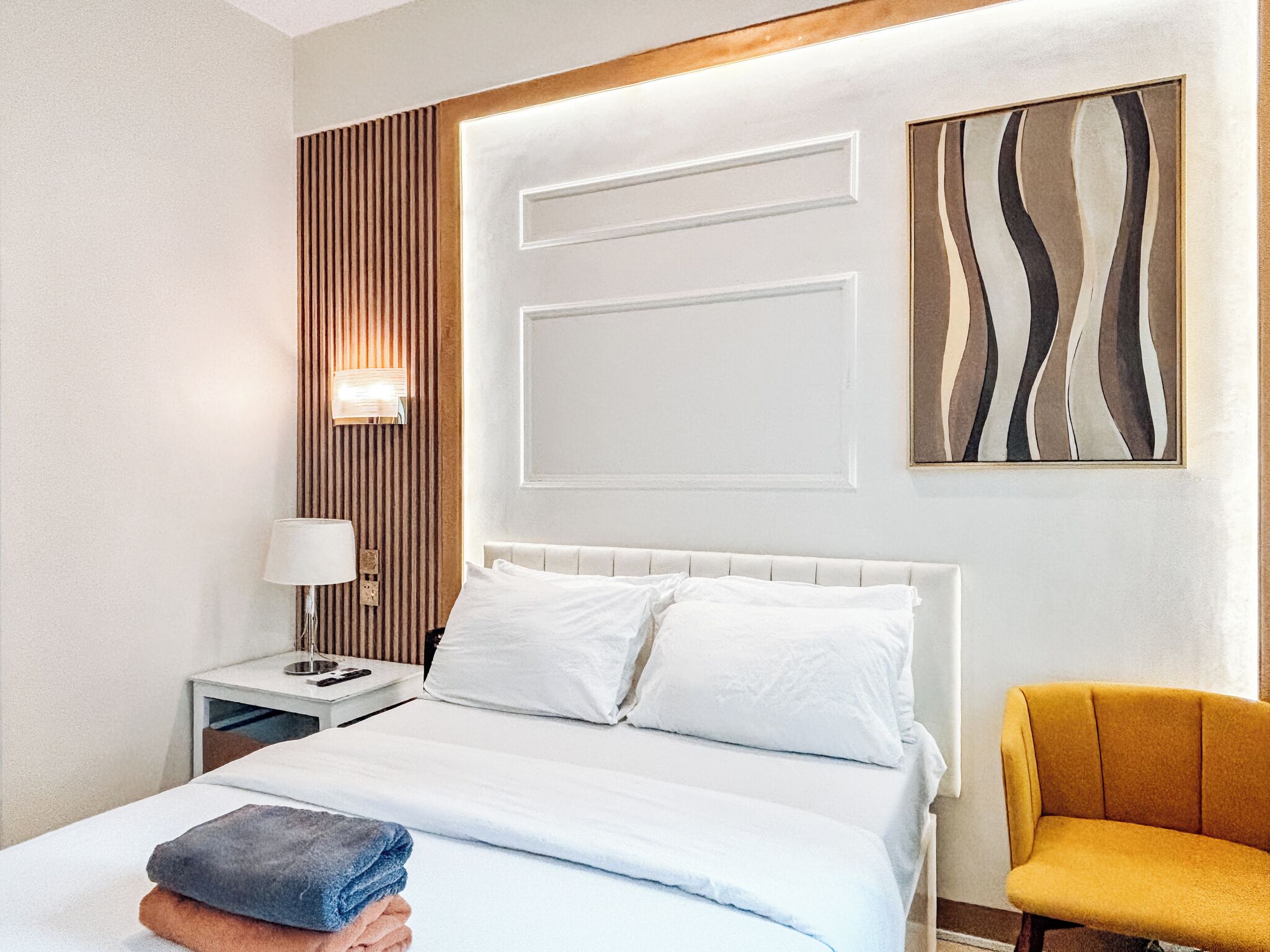 A hotel room with a bed, a bedside table with a lamp, a chair, and a wall-mounted artwork.