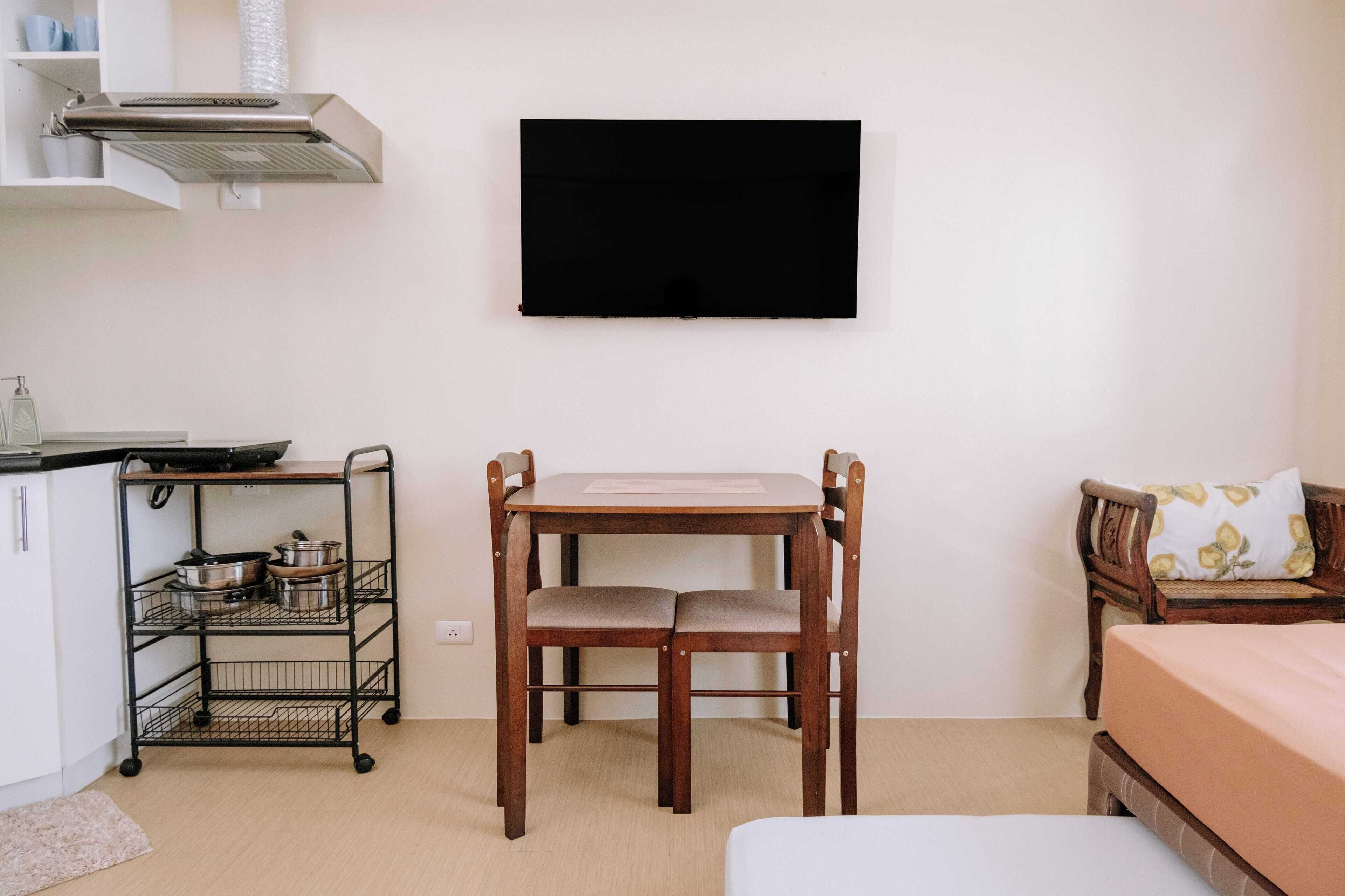 A compact room with a wooden table, two chairs, a bed, a TV mounted on the wall, a kitchenette with a stove and shelves, and a small rug.