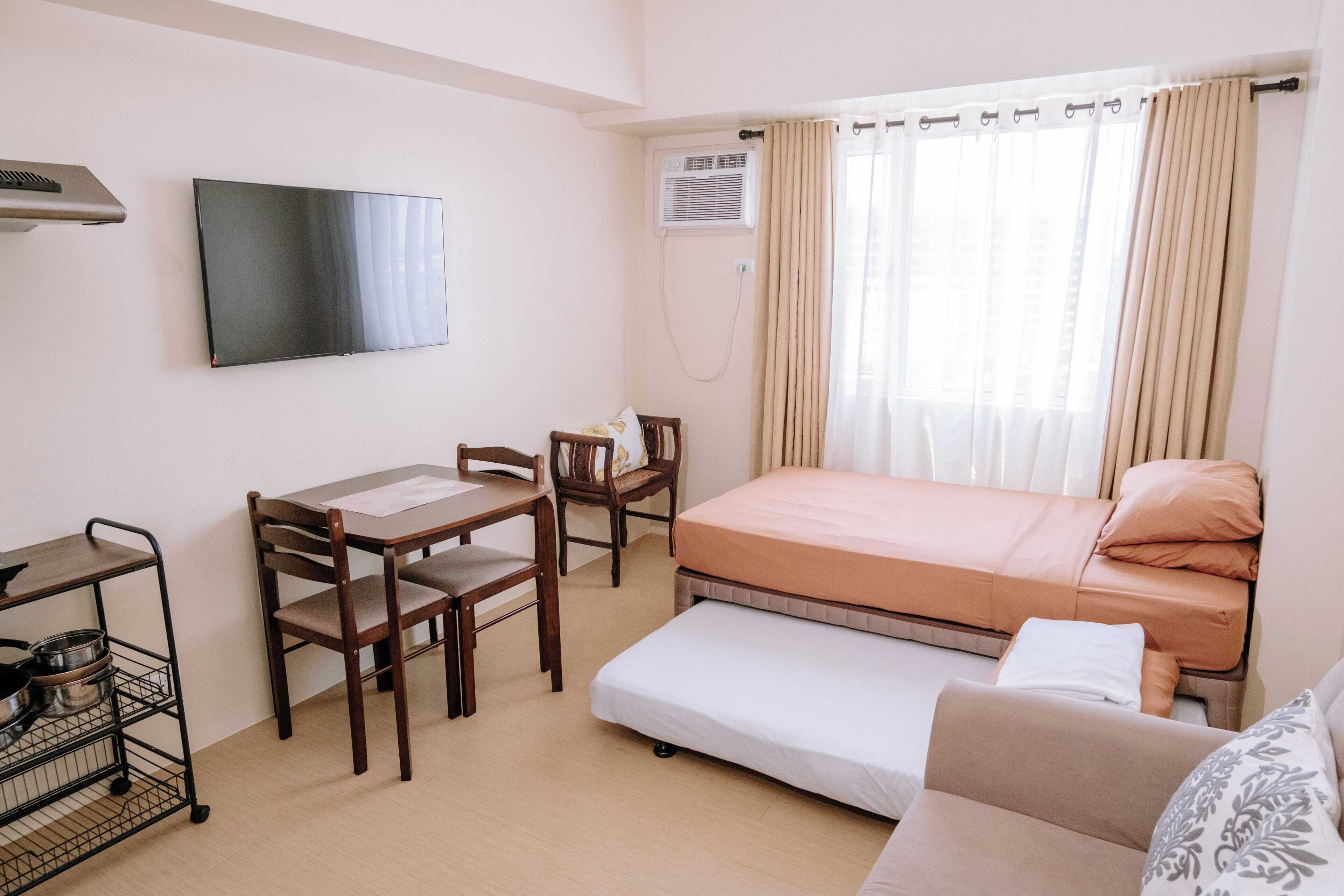 A compact room with a bed, a desk with chairs, a TV, and an air conditioning unit.