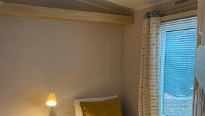 A bedroom with a bed, a bedside table with a lamp, a window with curtains, and a wooden headboard. - Cosy Grasmere Static Caravan (Natland)