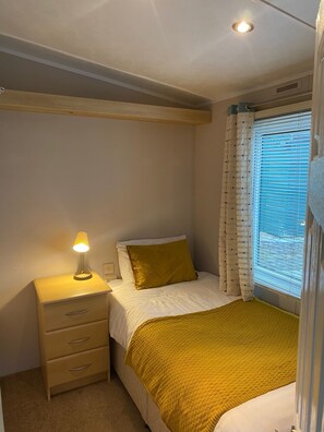 A bedroom with a bed, a bedside table with a lamp, a window with curtains, and a wooden headboard. - Cosy Grasmere Static Caravan (Natland)